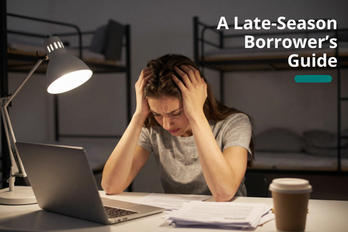 A young woman sits at a desk illuminated by a desk lamp, holding her head in her hands. She looks down at a pile of papers in front of her. An open laptop and a paper coffee cup are in the foreground.