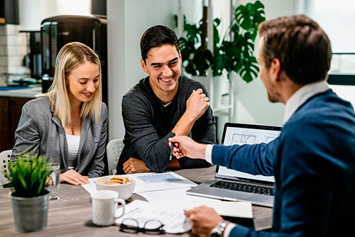 A man and woman sit across a desk from another man. He offers them a pen to sign paperwork.